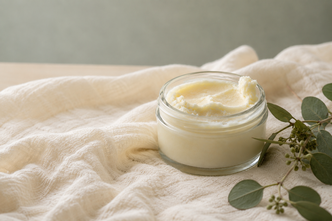 An open glass jar of ivory beef tallow balm on a soft folded cream muslin baby blanket with a sprig of greenery, a gentle fragrance-free moisturizer for a baby's dry skin.